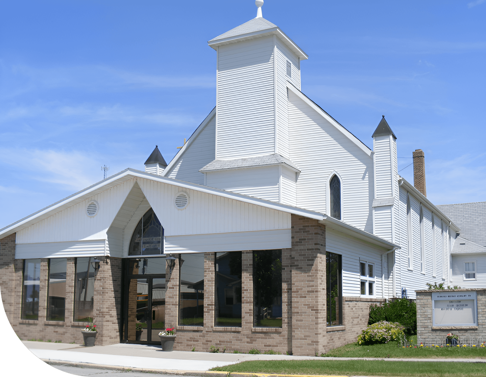 White church building with a brick base, featuring a prominent tower and an entrance with large glass windows. The sky is clear and blue.